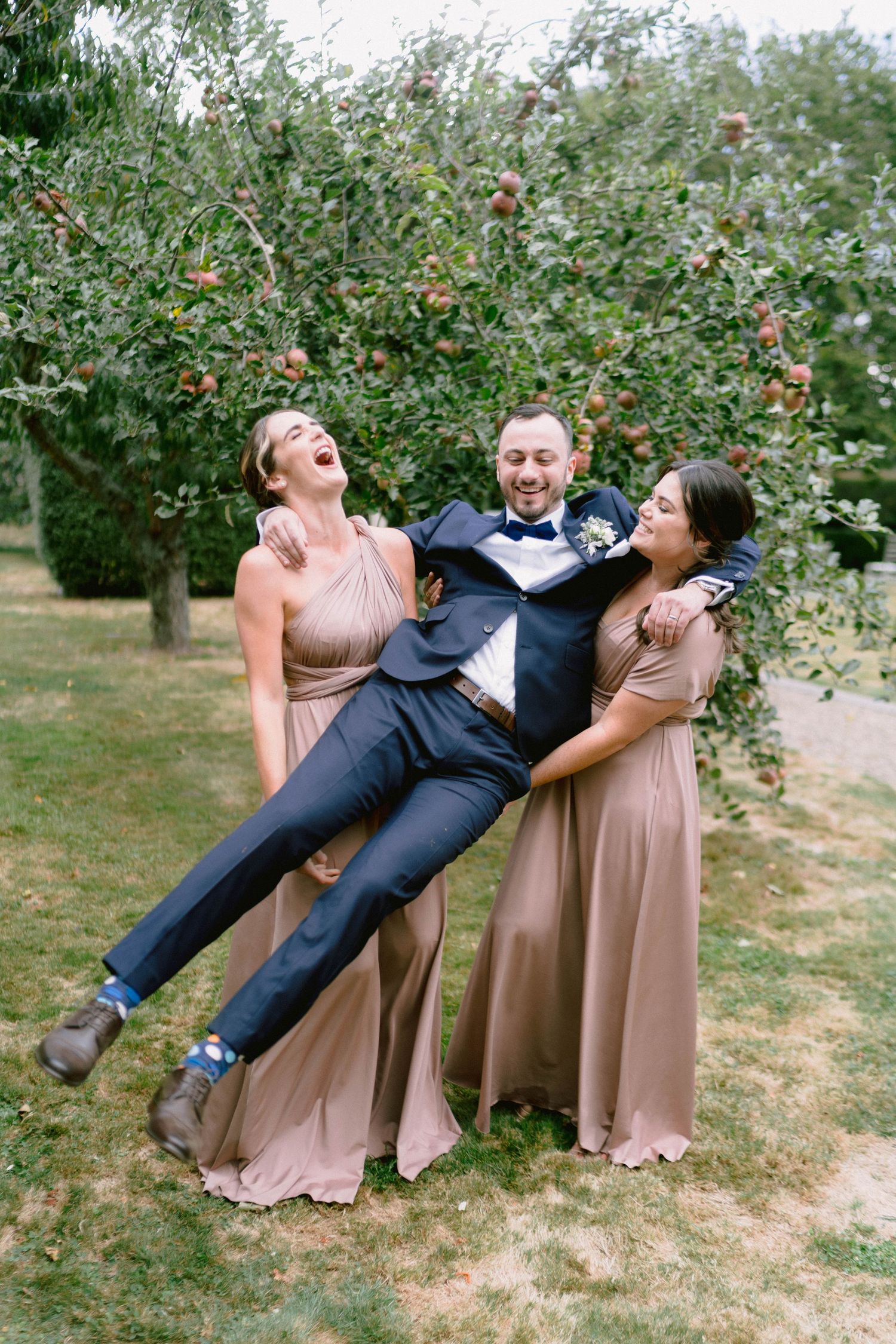 Wedding attendants in blush dresses playfully lift a guest in a navy suit at an orchard.