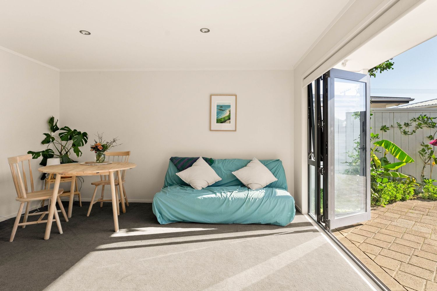 Bright studio apartment interior with teal sofa, dining area, and bi-fold doors opening to patio.