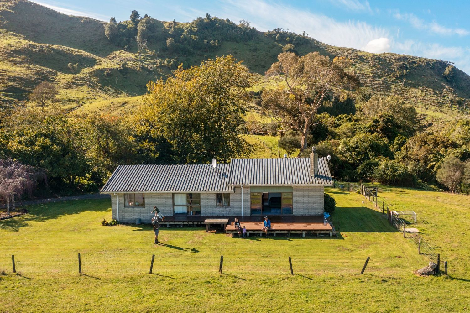 White rural farmhouse with metal roof sits on green hillside surrounded by trees and mountains in New Zealand countryside.
