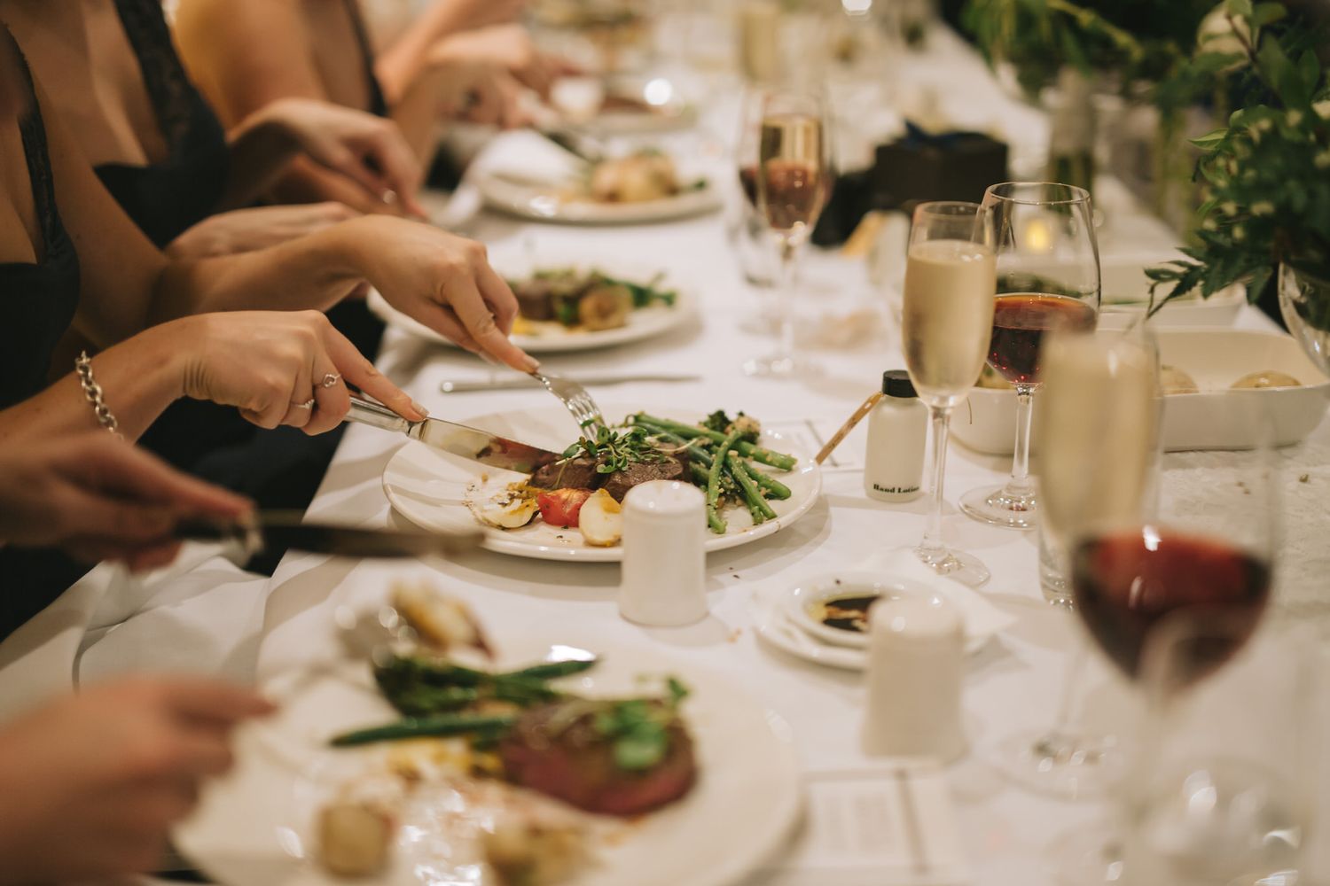 Close-up view of dining guests hands using cutlery at an elegant table setting with wine glasses and plates.
