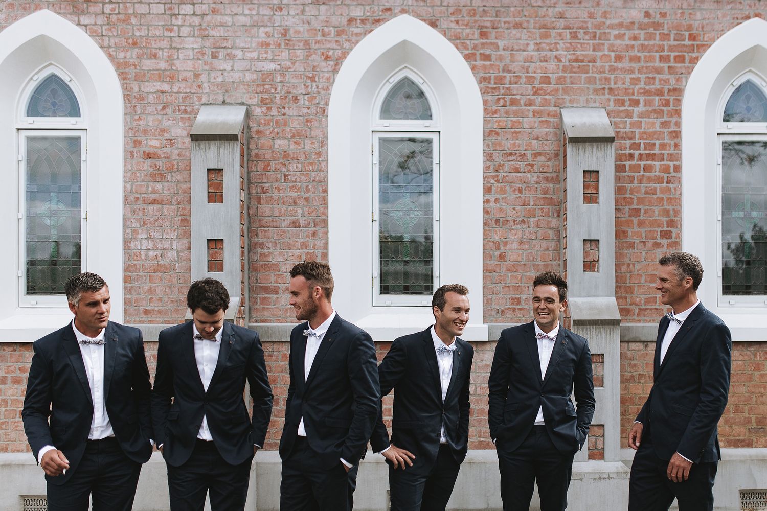 Groomsmen dressed in black suits stand together against a brick wall with white gothic arched windows.
