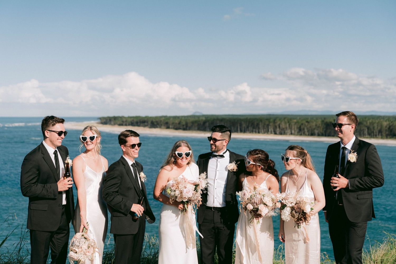 Wedding party lined up outdoors against scenic ocean and forest backdrop in formal attire.
