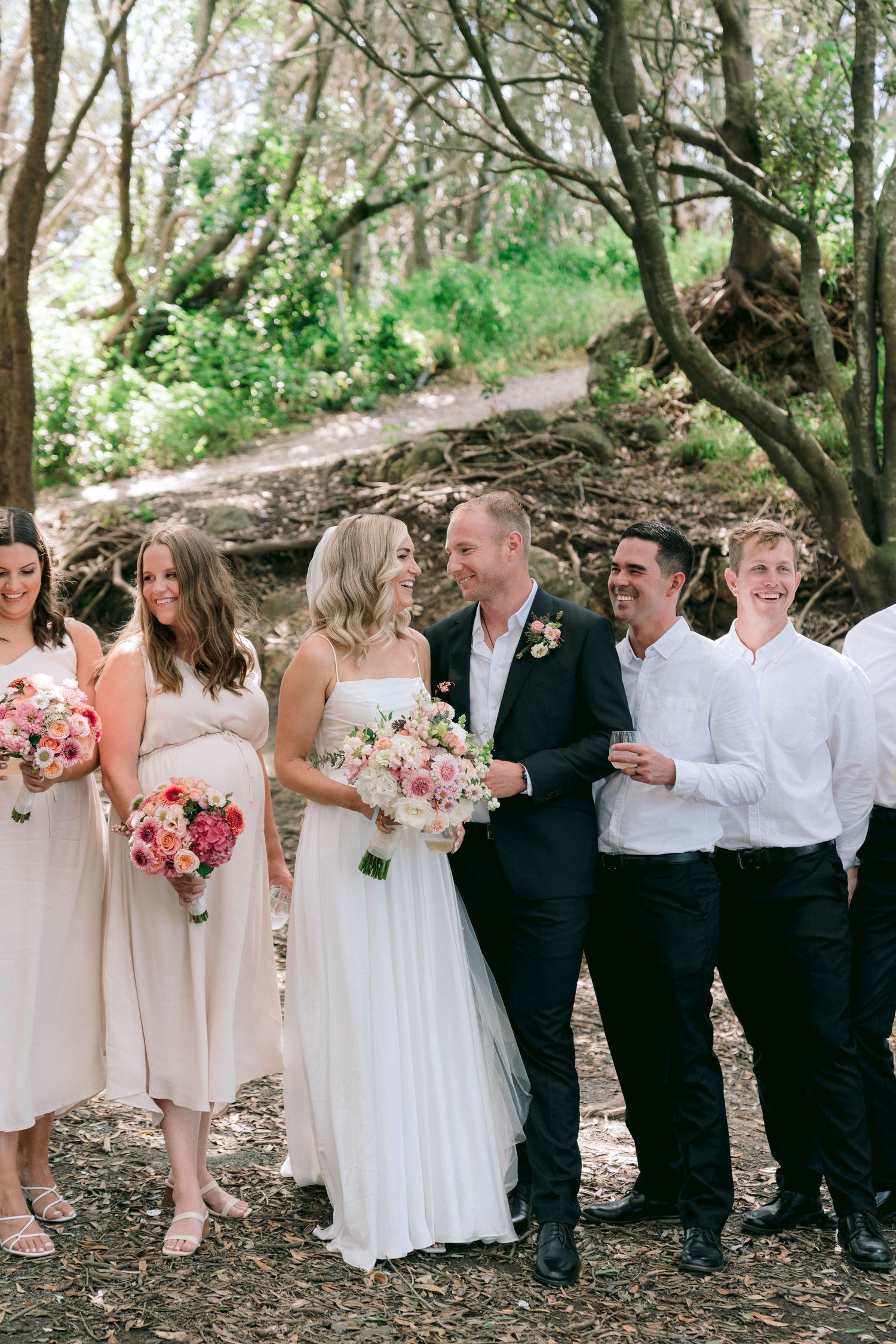 Wedding party stands on a wooded path with bridesmaids in blush dresses holding pink and white floral bouquets.