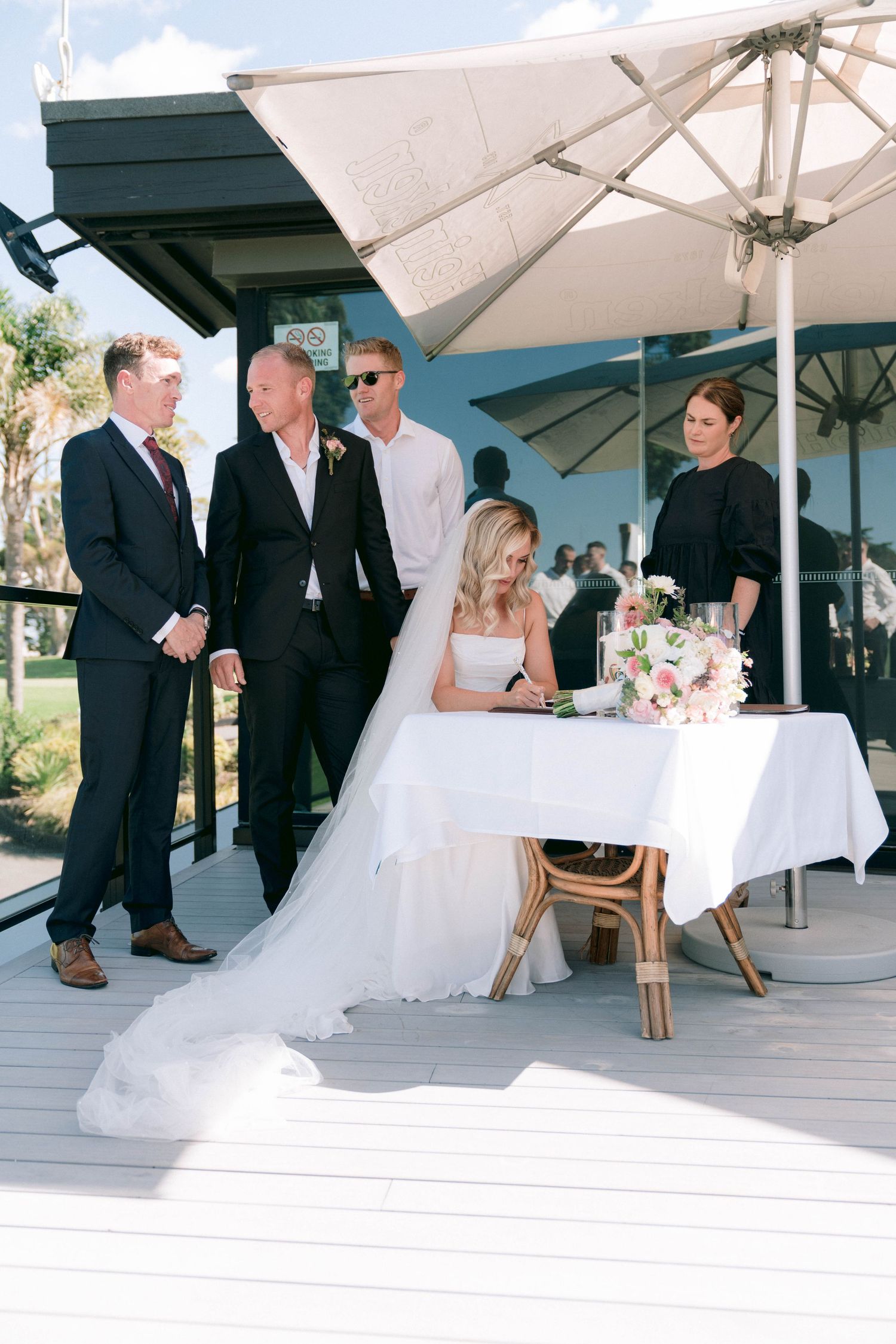 A wedding signing ceremony takes place on an outdoor deck under a white umbrella with guests gathered around.