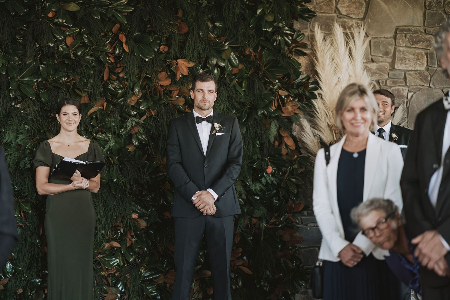 Wedding attendees stand together during an outdoor ceremony against a stone wall and foliage backdrop.
