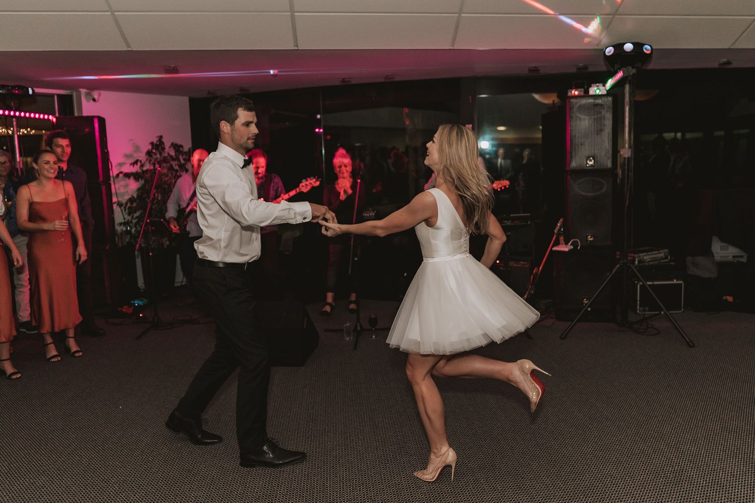 Wedding guests dance together on a dimly lit dance floor with pink lighting creating a festive party atmosphere.