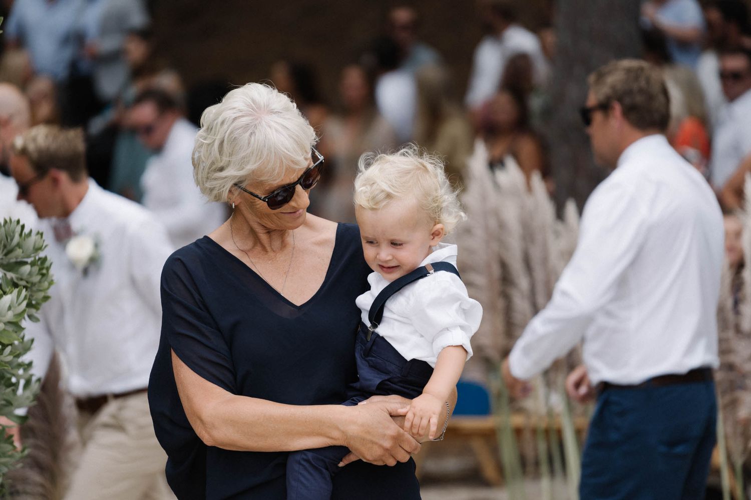 A person in sunglasses holds a small child wearing formal white attire at an outdoor gathering.