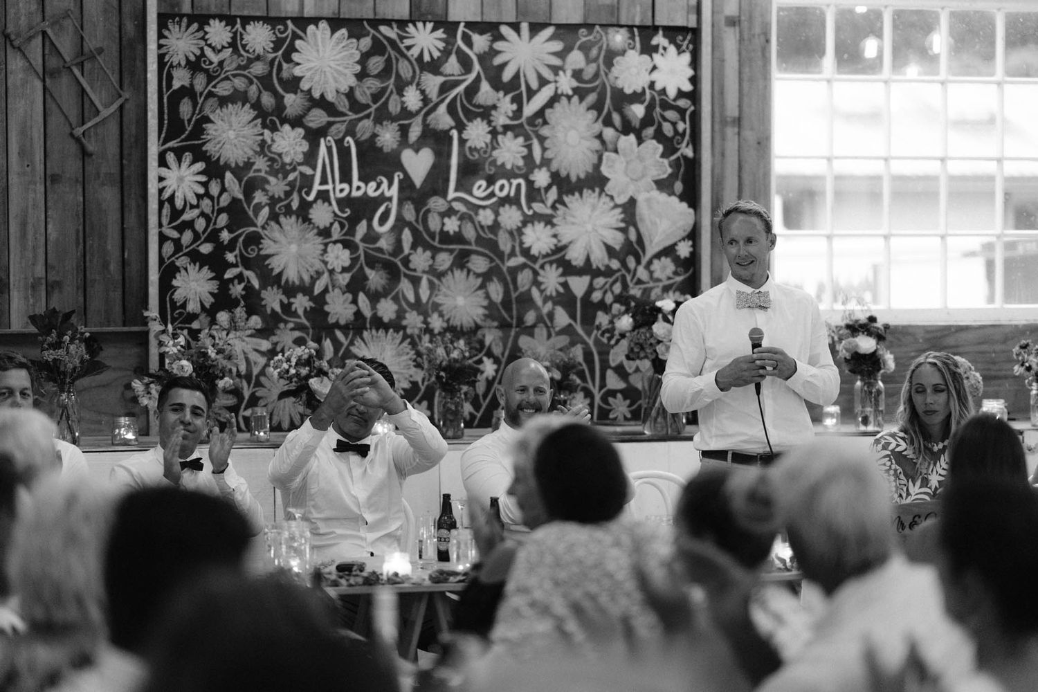 Black and white photo of people gathered at a dining event with decorative floral wallpaper in the background.