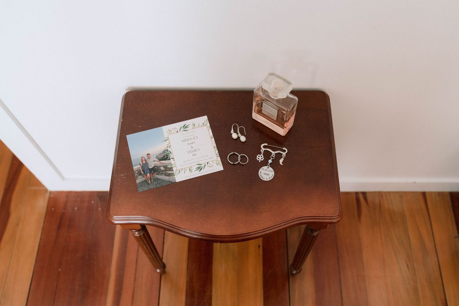 A vintage wooden side table displays a postcard, ring, and small glass jar on its surface against a white wall and wood floor.