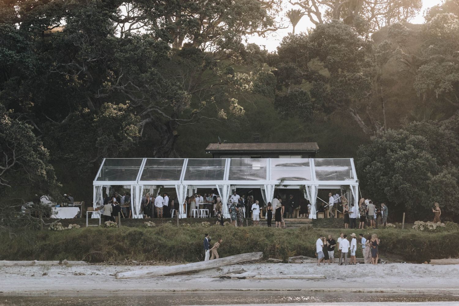 Clear tent wedding venue setup on a beach with mountains and forest in background at dusk.