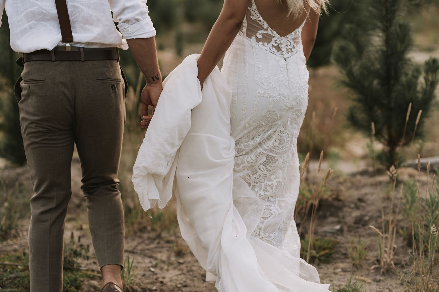 Close-up view of bride in fitted lace wedding gown with train standing next to groom in brown dress pants outdoors.