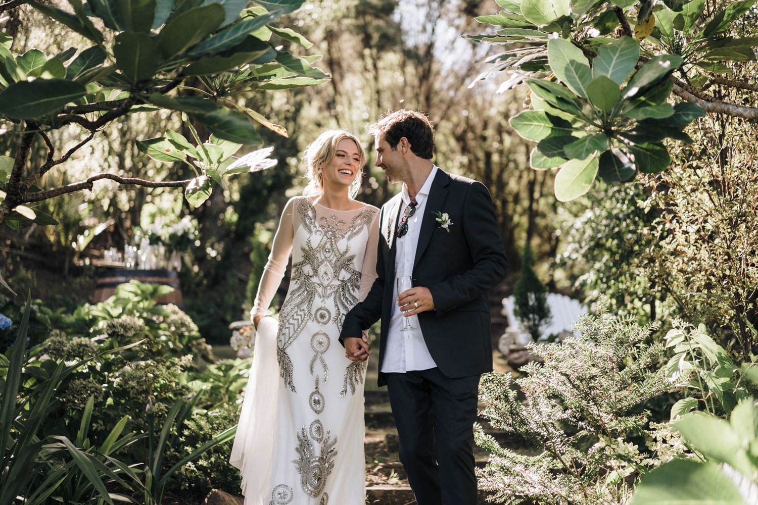 Newlywed couple walking together through a lush tropical garden in elegant wedding attire.