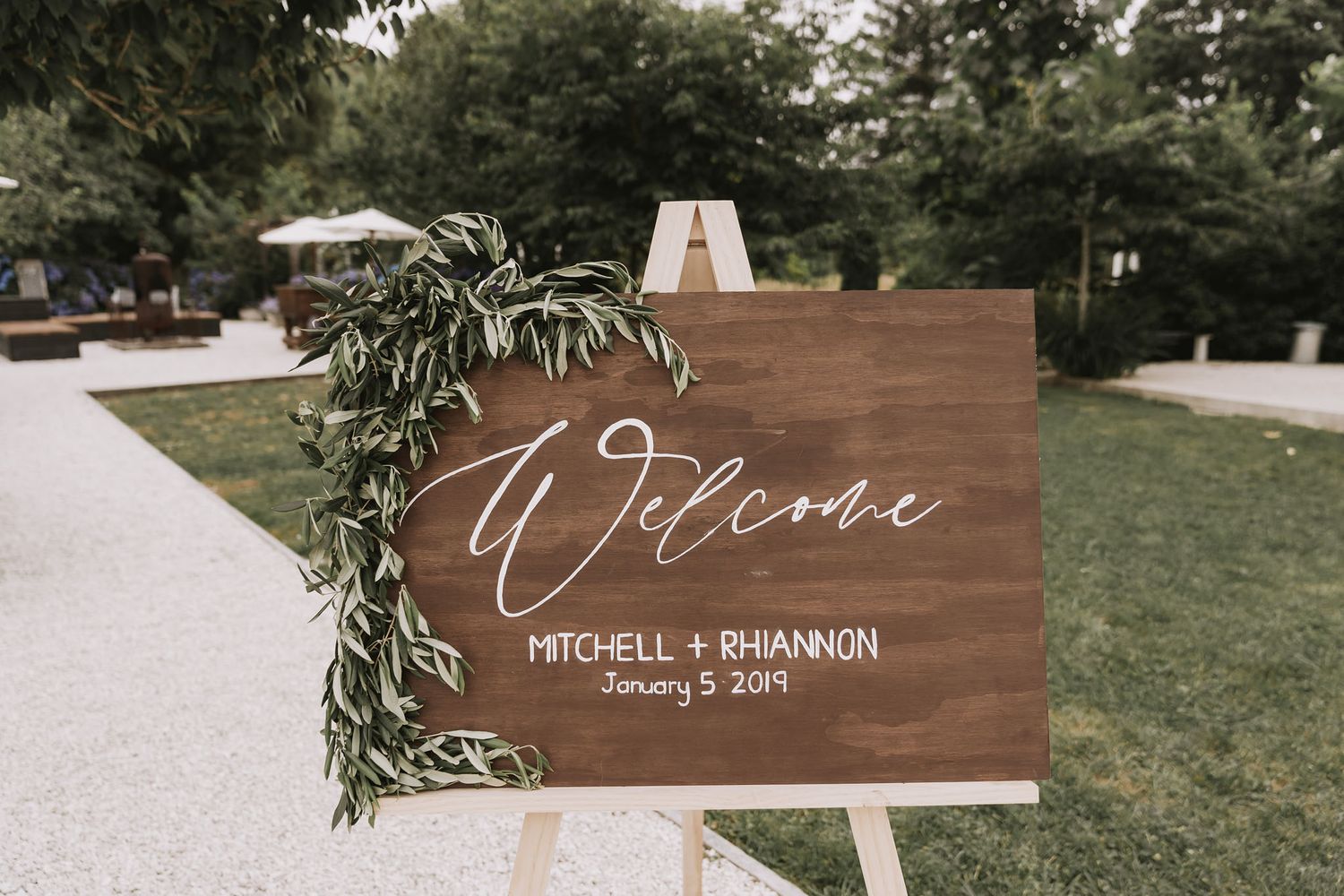 Wooden welcome sign with elegant white calligraphy and greenery decoration at outdoor wedding ceremony.