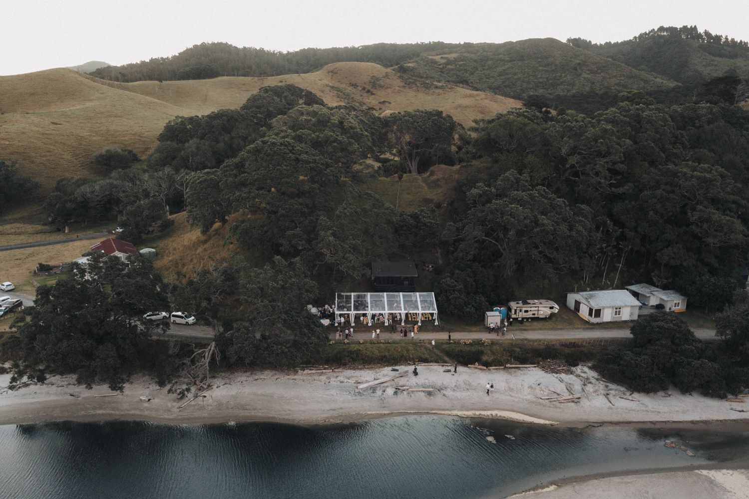 Aerial view of beachfront houses and buildings nestled among trees on a curved coastline with rolling hills in background.