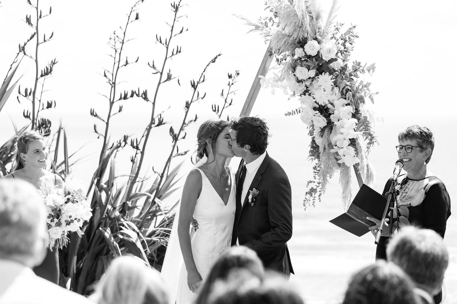 A row of wedding guests at an outdoor ceremony stand together holding programs and smiling on a sunny day.