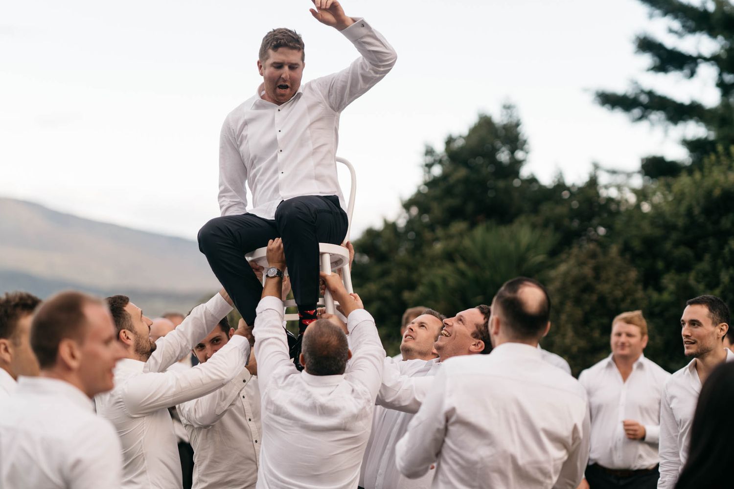A group of groomsmen in white shirts lift a person in celebration during an outdoor wedding event.