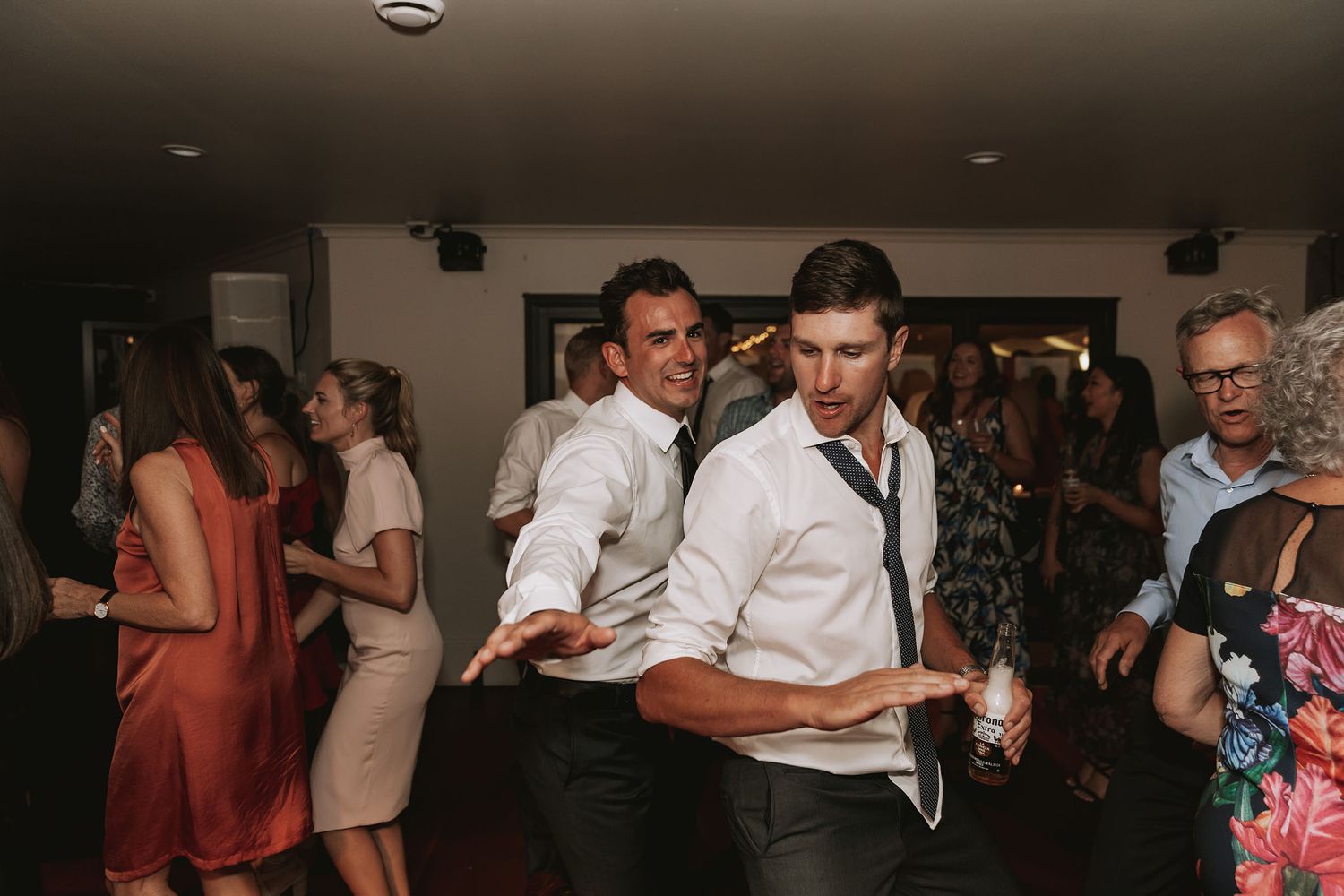People dancing and enjoying themselves at a wedding reception in a dimly lit venue.