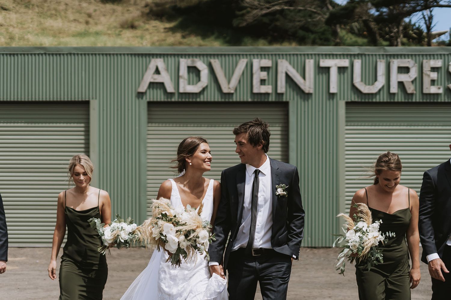 Wedding party walks past green industrial building with Adventure sign, bride in white dress holds floral bouquet.