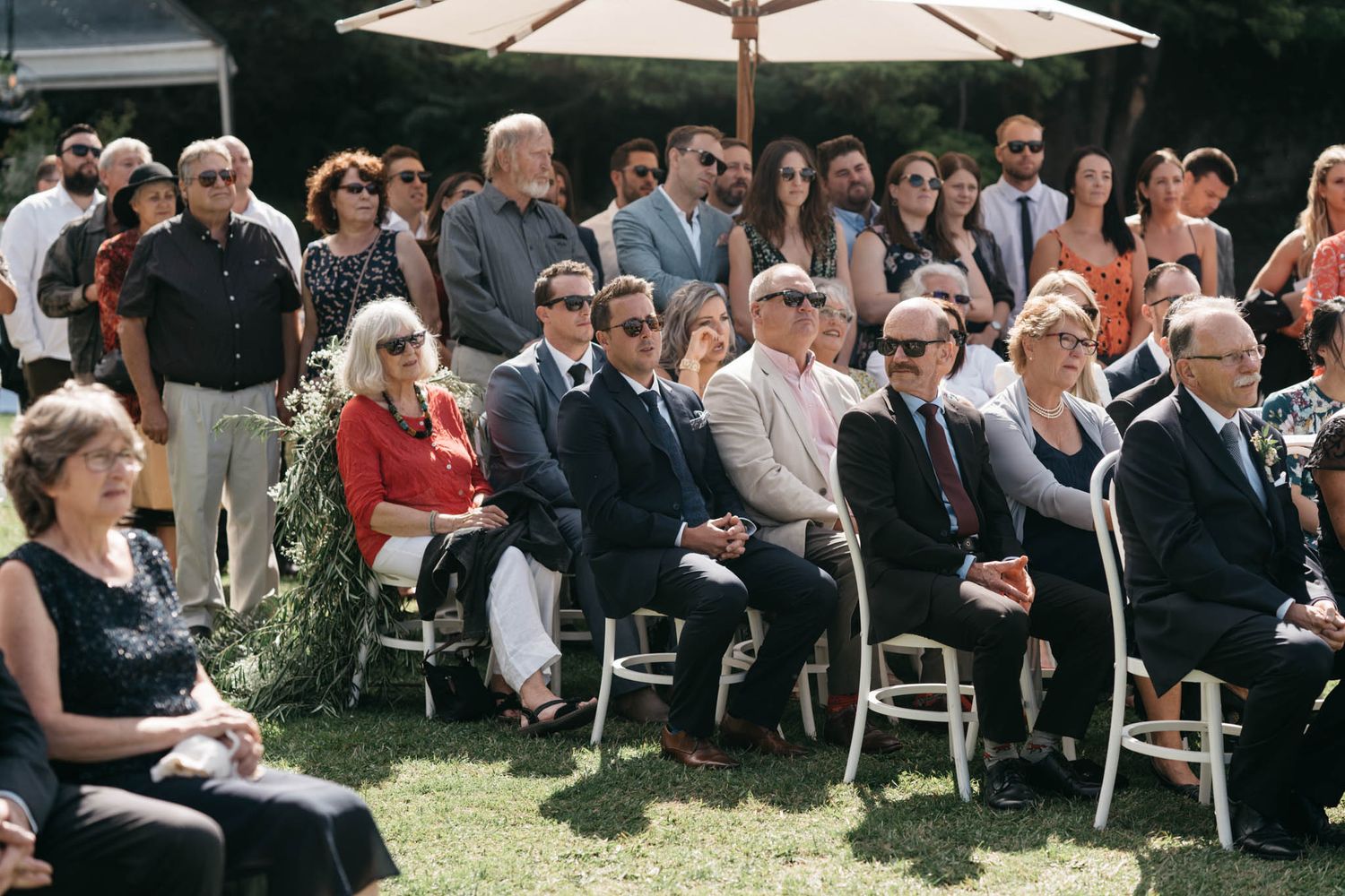 Wedding guests seated in white chairs under outdoor umbrellas watch a ceremony on a sunny afternoon.
