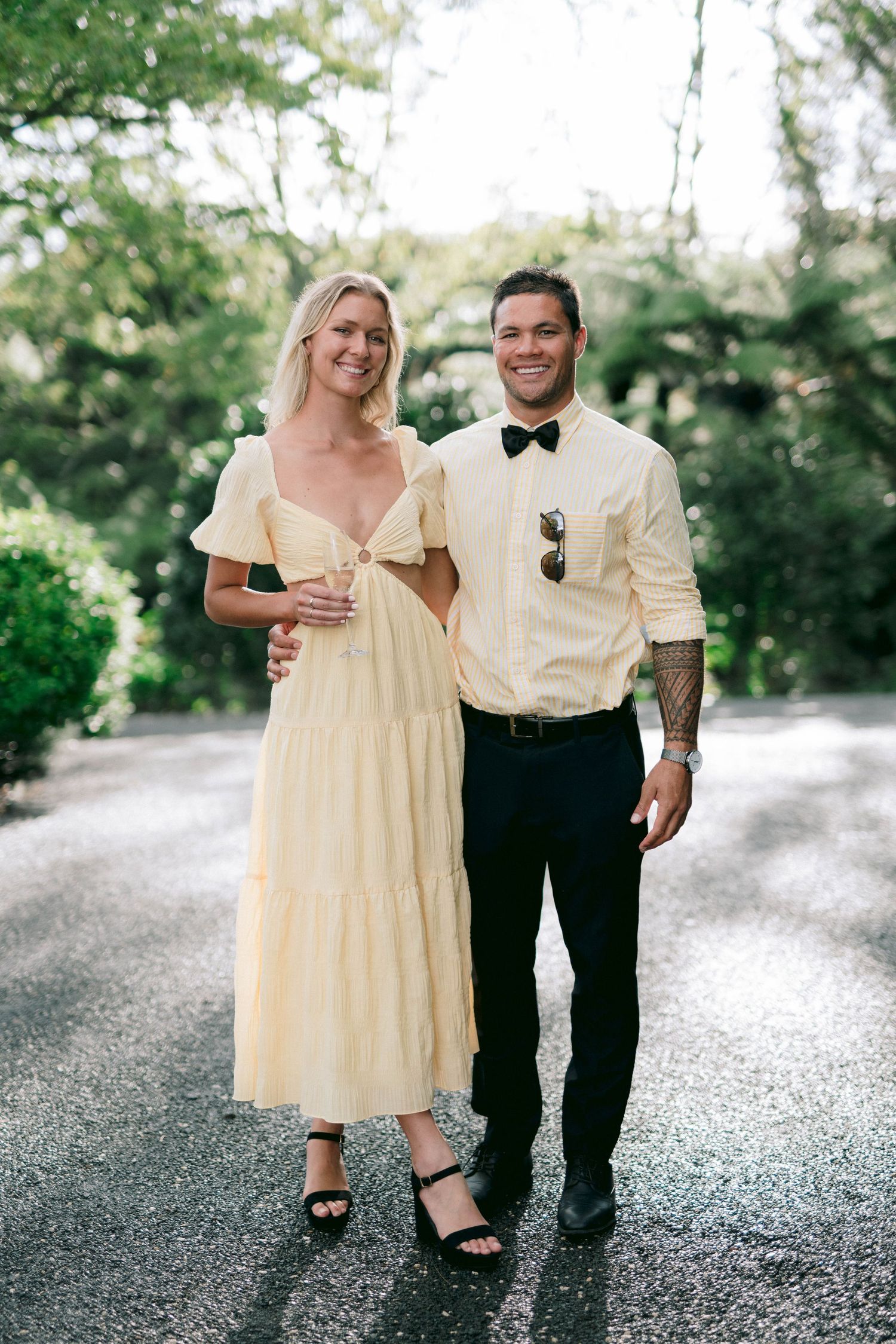 A couple in coordinated cream and white outfits smiles while standing on a path surrounded by trees.