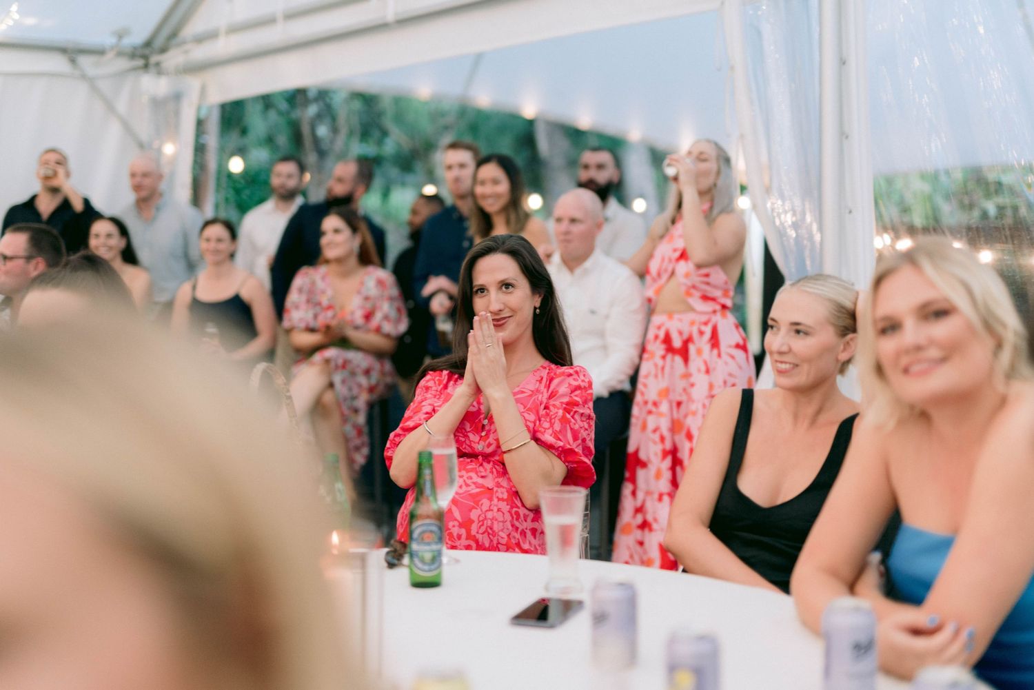 Wedding reception guests in bright attire react with excitement during a celebration under a white tent.