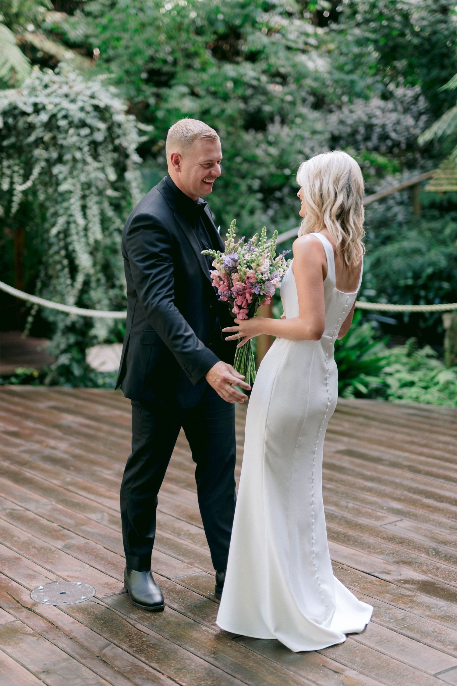 A couple shares a joyful moment on a wooden deck surrounded by lush greenery with a wedding bouquet.