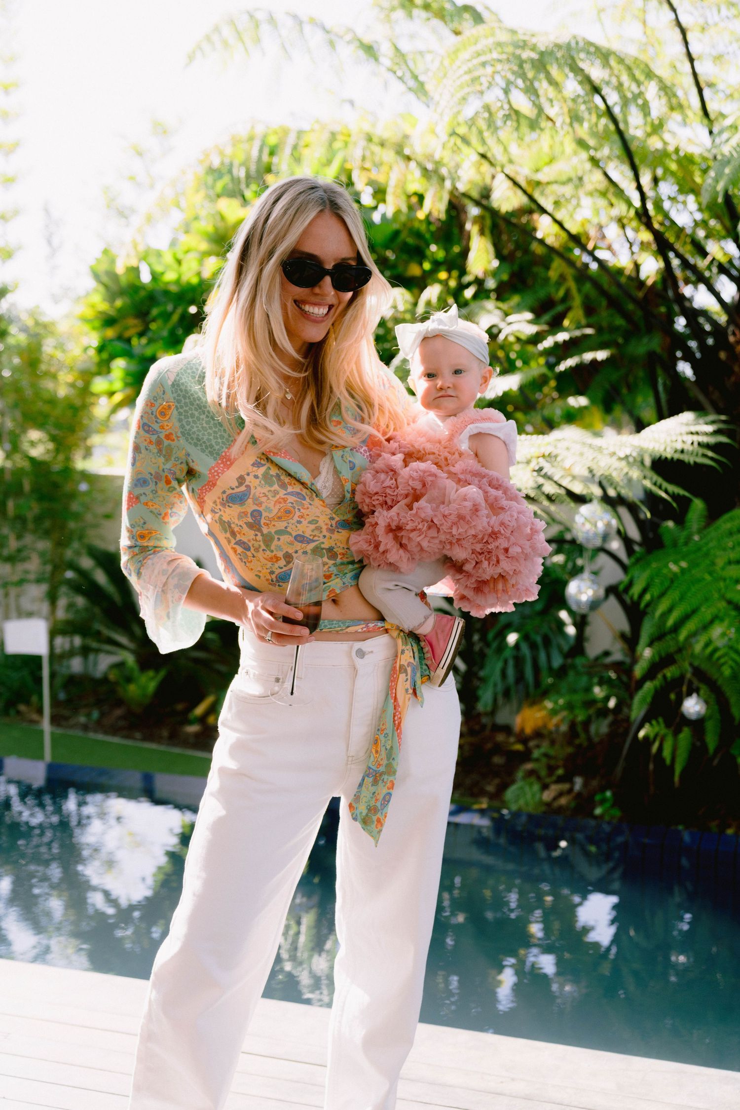 Adult holding baby in pink tulle dress near pool surrounded by tropical plants.