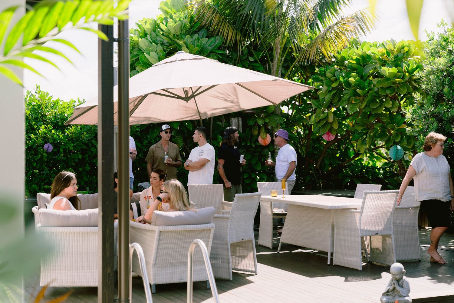 Guests relax in white chairs on an outdoor patio surrounded by lush tropical plants and umbrellas.