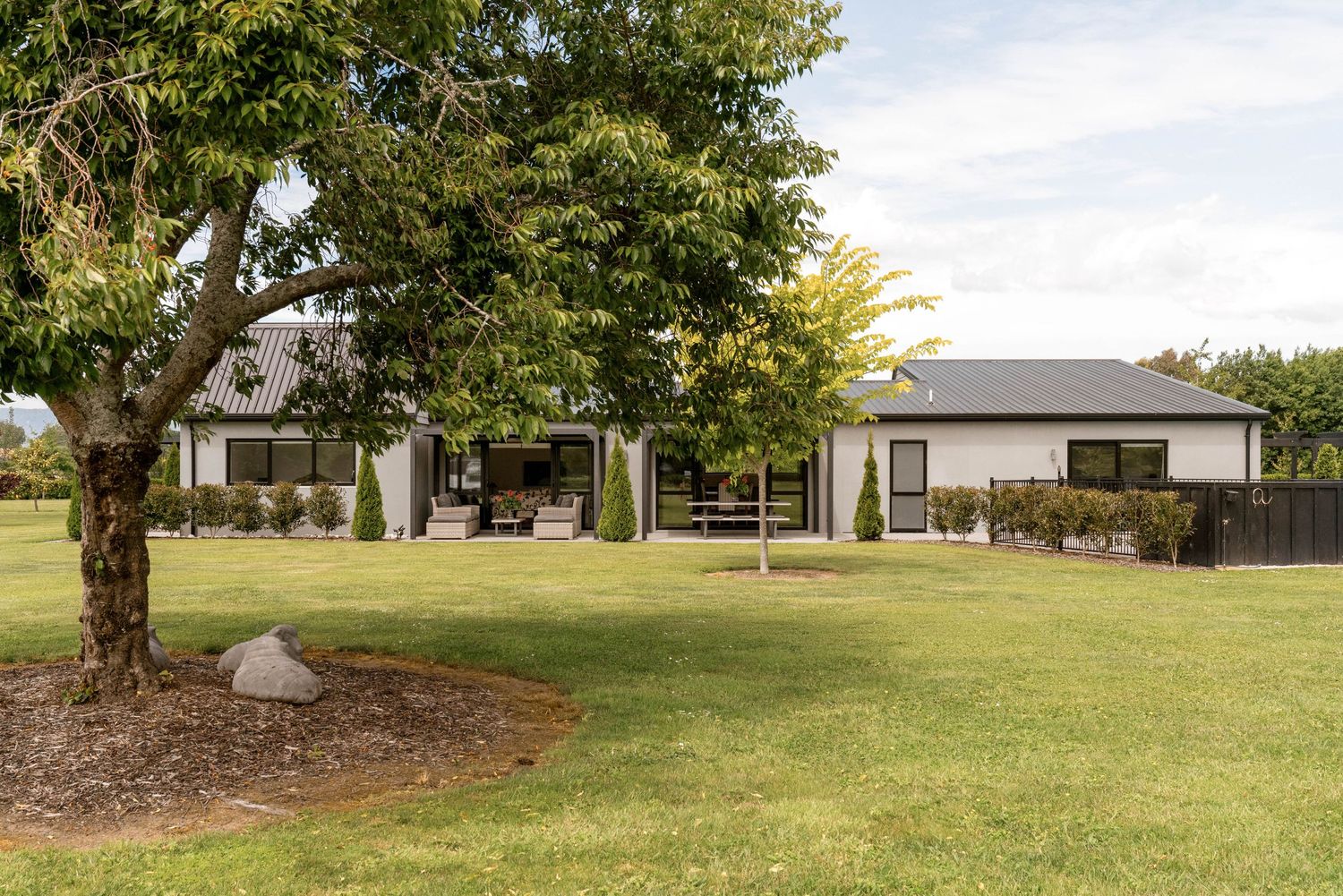 Modern single-story ranch house with white exterior and lush green lawn surrounded by mature trees on spacious property.