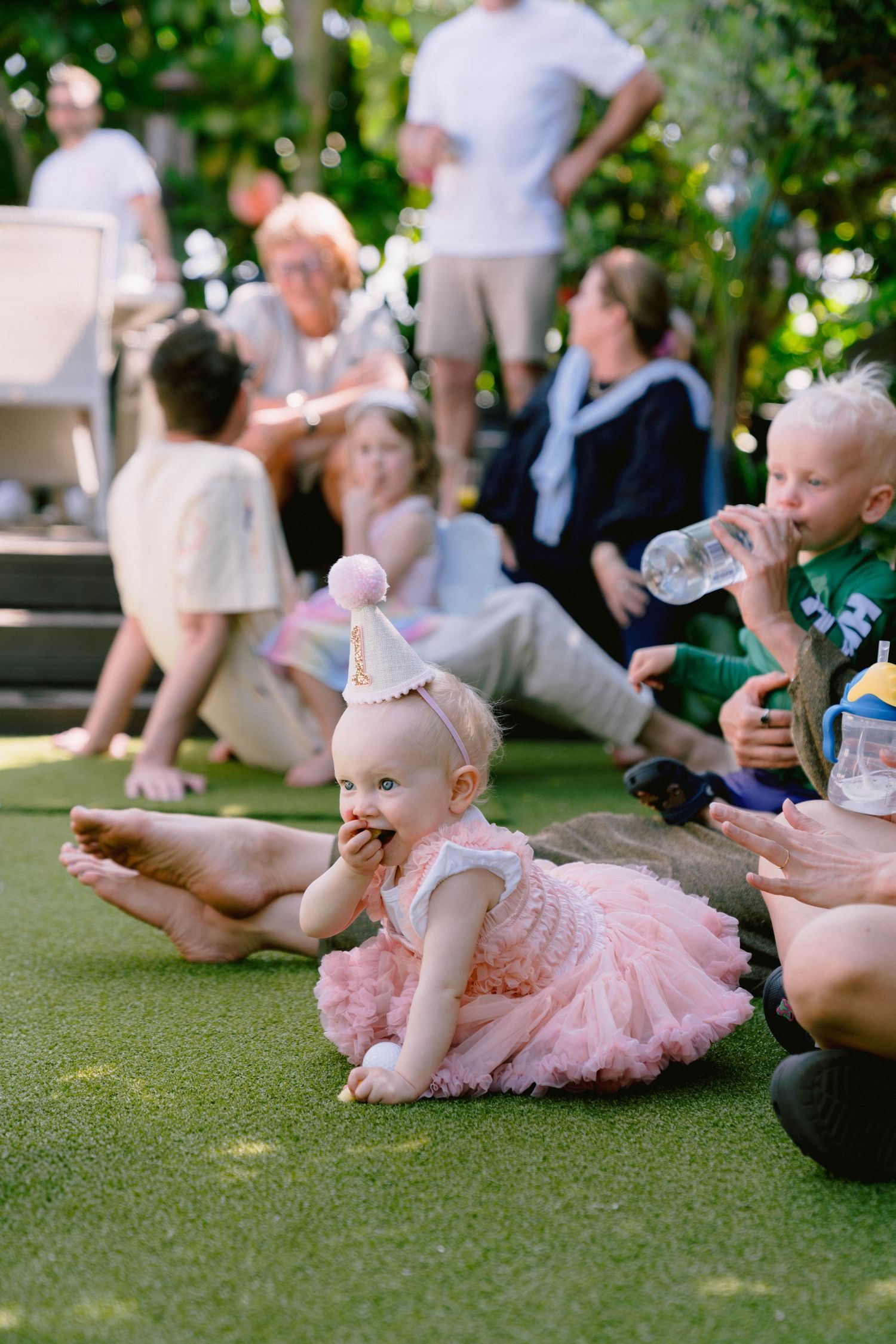 Baby wearing pink tutu dress and party hat sits on grass with people gathered at outdoor celebration.