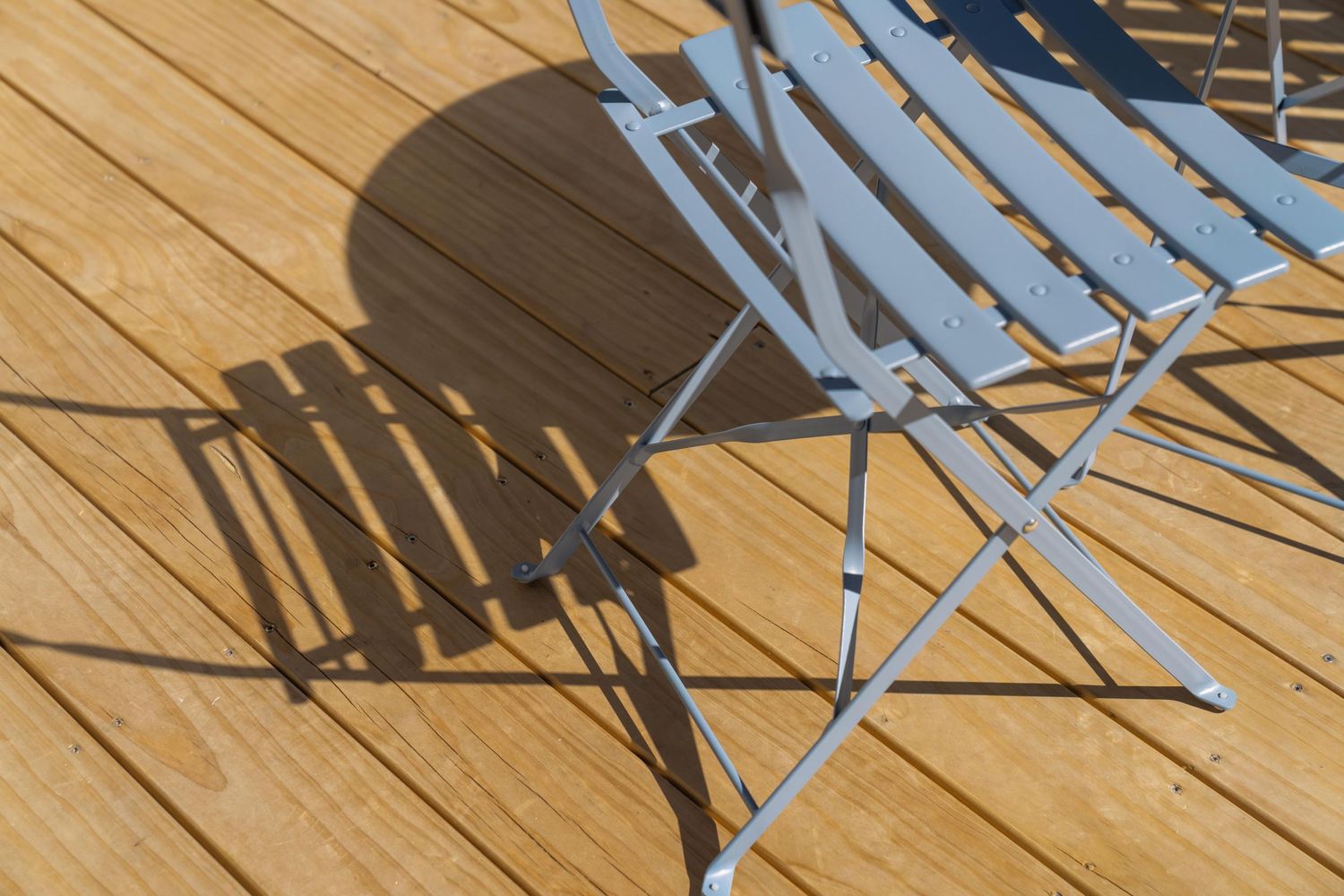 Metal folding chair casting dramatic shadows on wooden deck planks in bright sunlight.