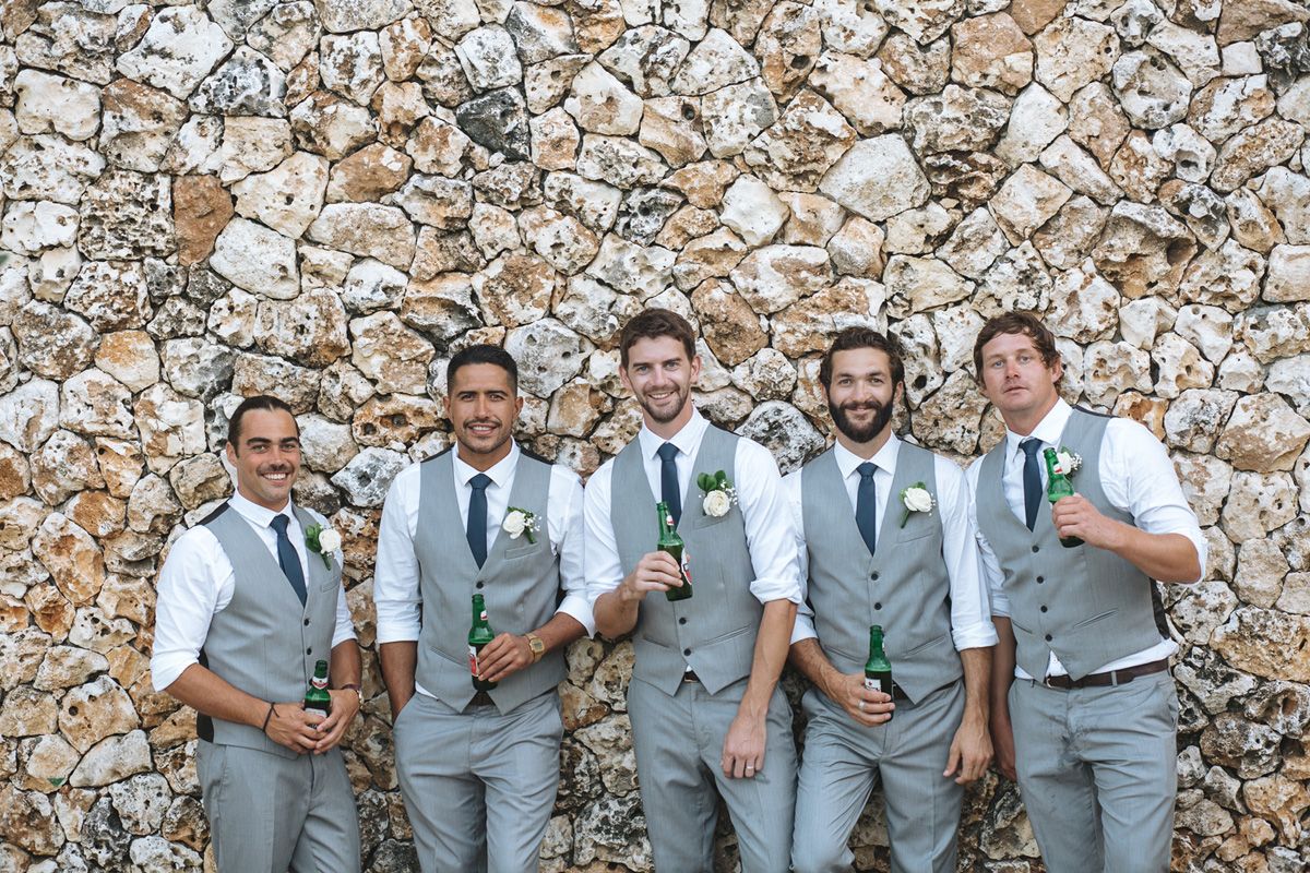 Groomsmen in light gray suits pose together against a stone wall backdrop.
