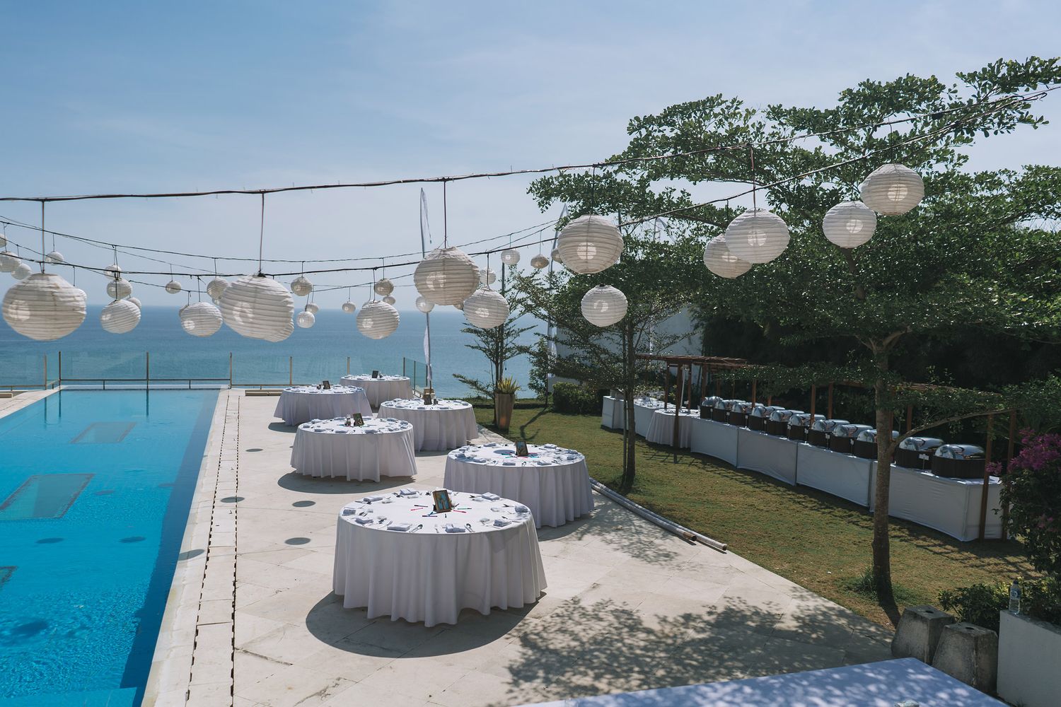 Outdoor wedding reception setup with white tables and lanterns overlooking infinity pool and ocean view.
