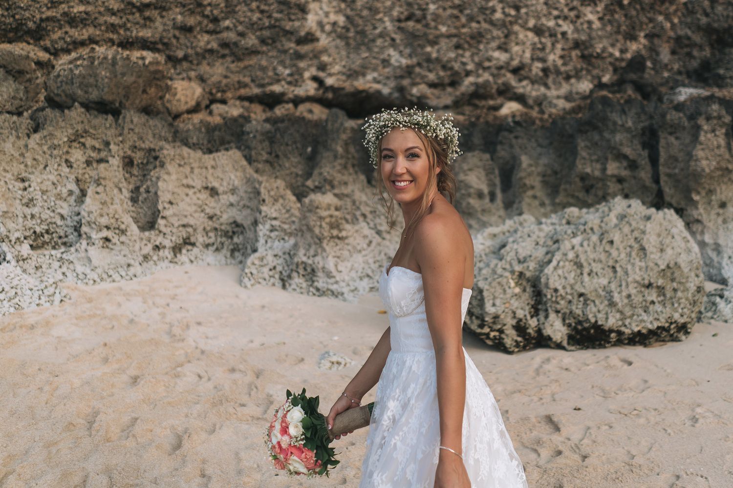 Bride in white dress holding bouquet poses against rocky beach landscape.