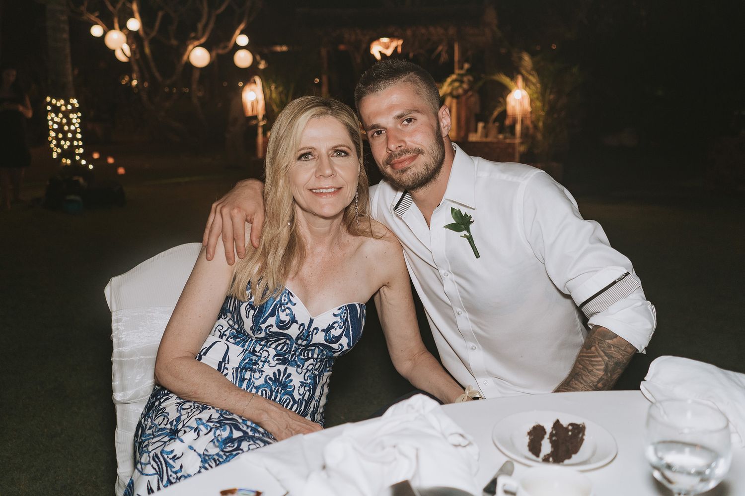 A couple poses together at a reception dinner table with decorative lighting in the background.