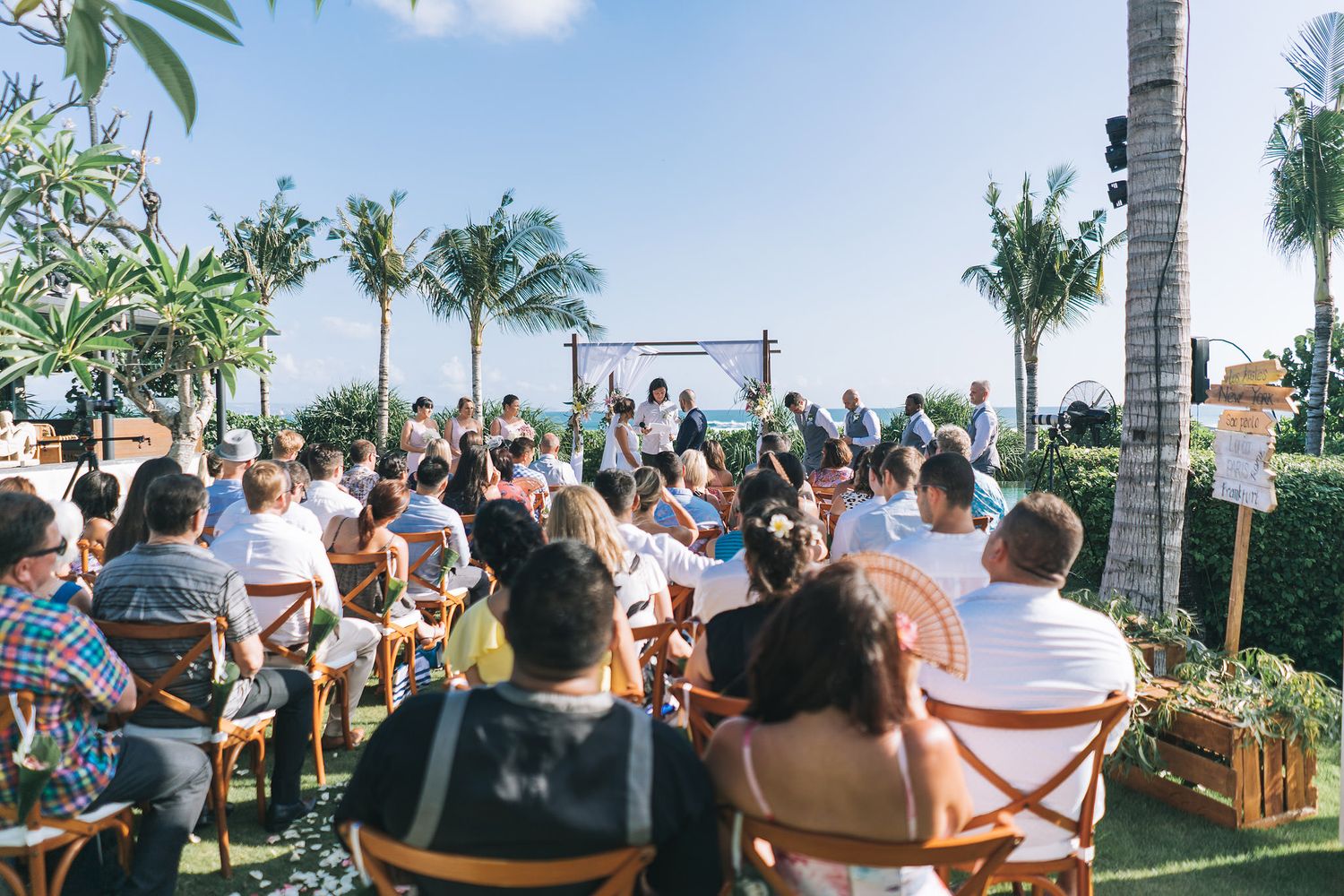Outdoor rooftop wedding ceremony with guests seated in wooden chairs surrounded by palm trees against a blue sky.