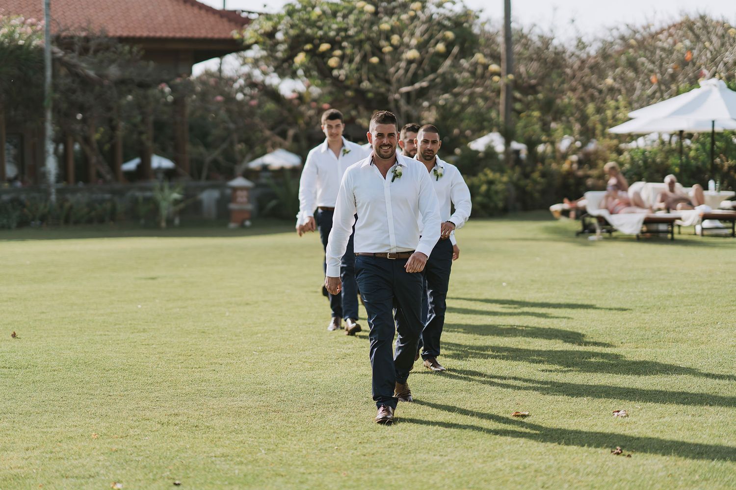 Groomsmen walking in formation across a manicured lawn at an outdoor event.