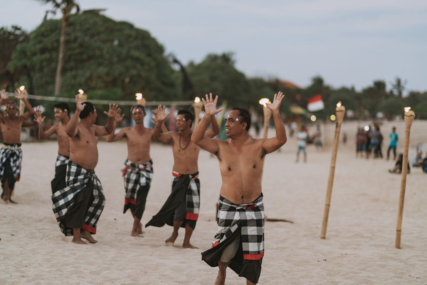 Group of performers in traditional sarongs practicing a ceremonial dance routine on a sandy beach.