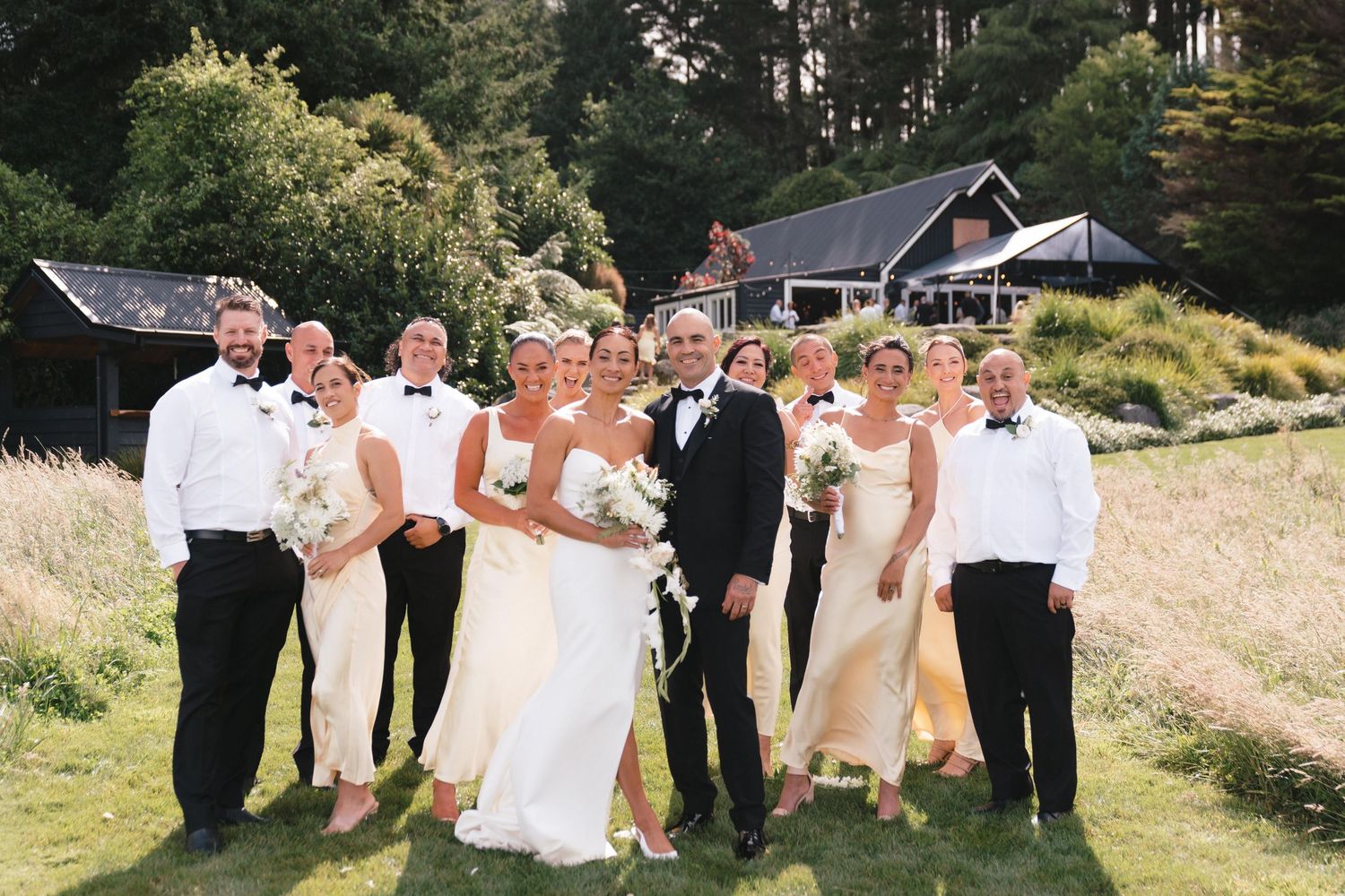 Wedding party stands in front of rustic cabins on a grassy hillside in natural outdoor setting.