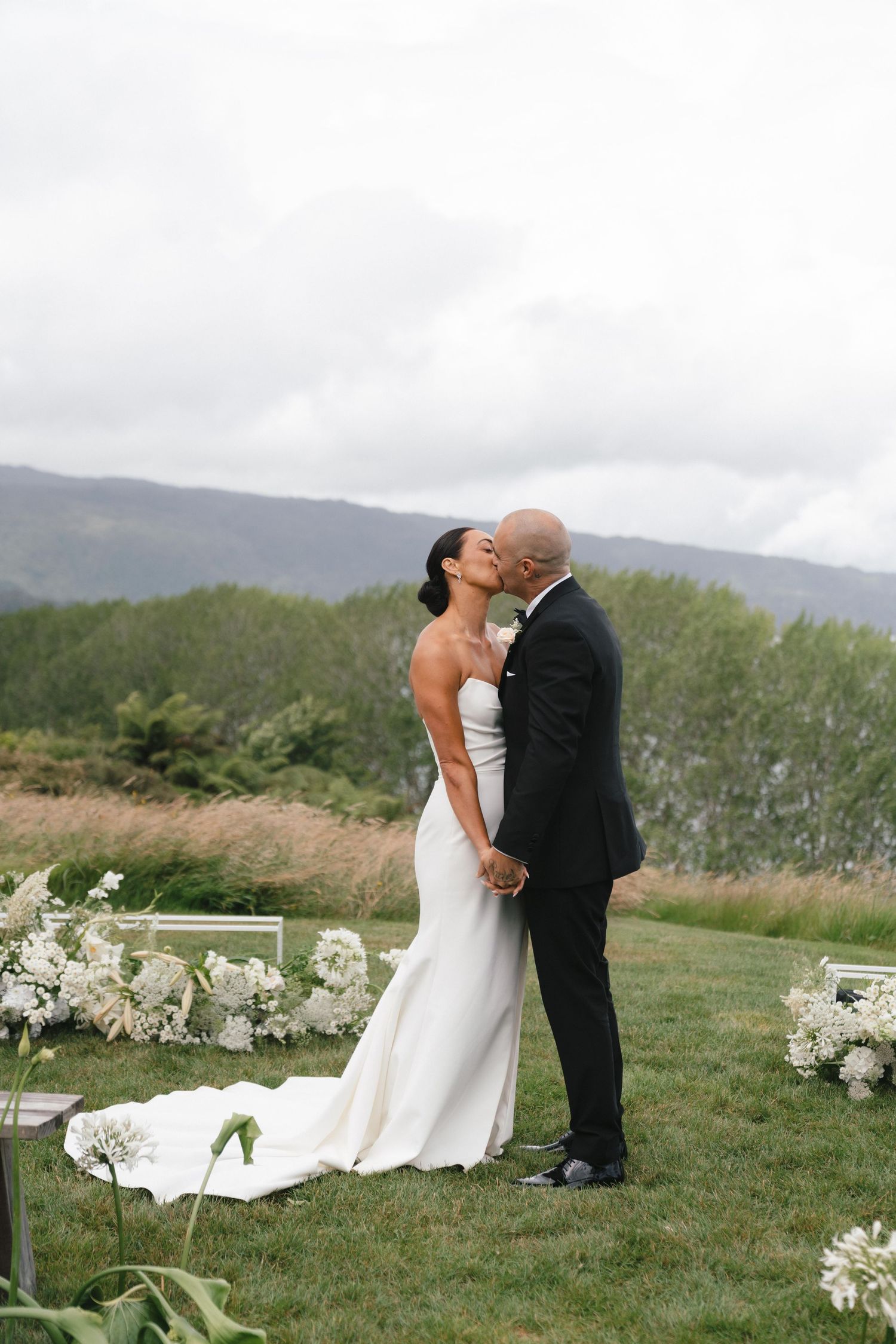 Newlyweds share a romantic moment on a hilltop with mountain views and white floral decorations.