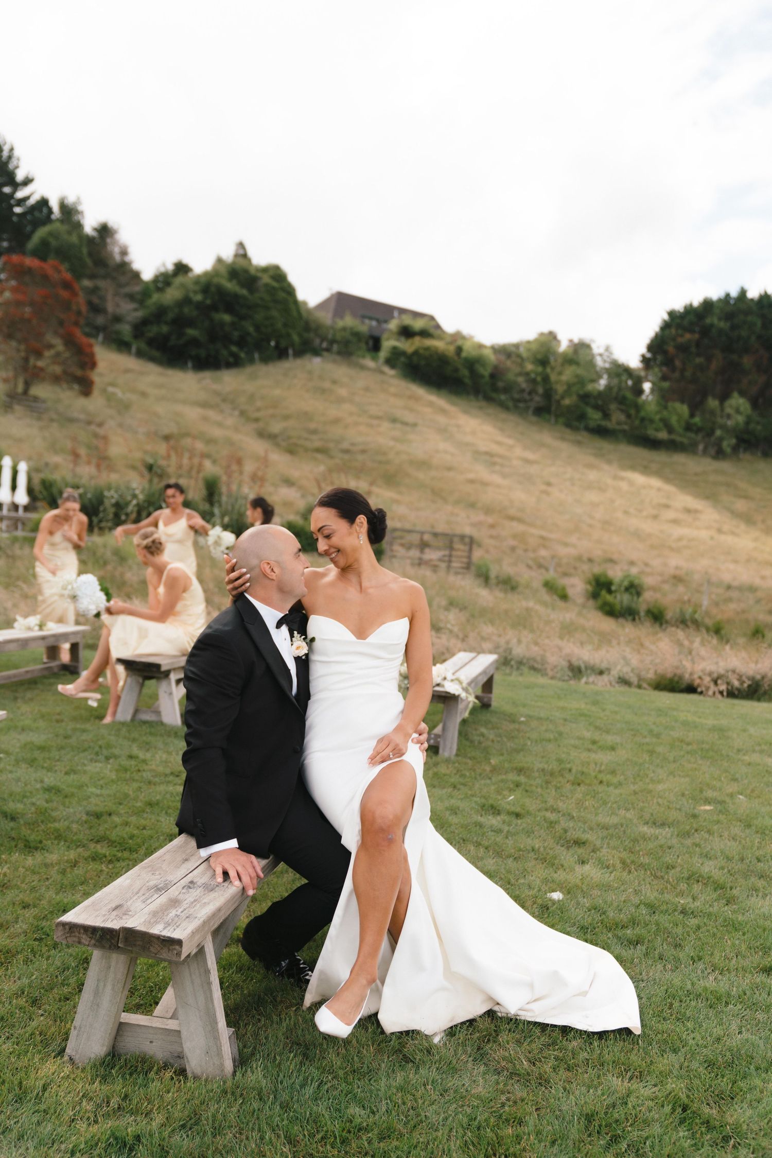 A couple in formal wedding attire sits together on a rustic wooden bench at an outdoor hillside venue.