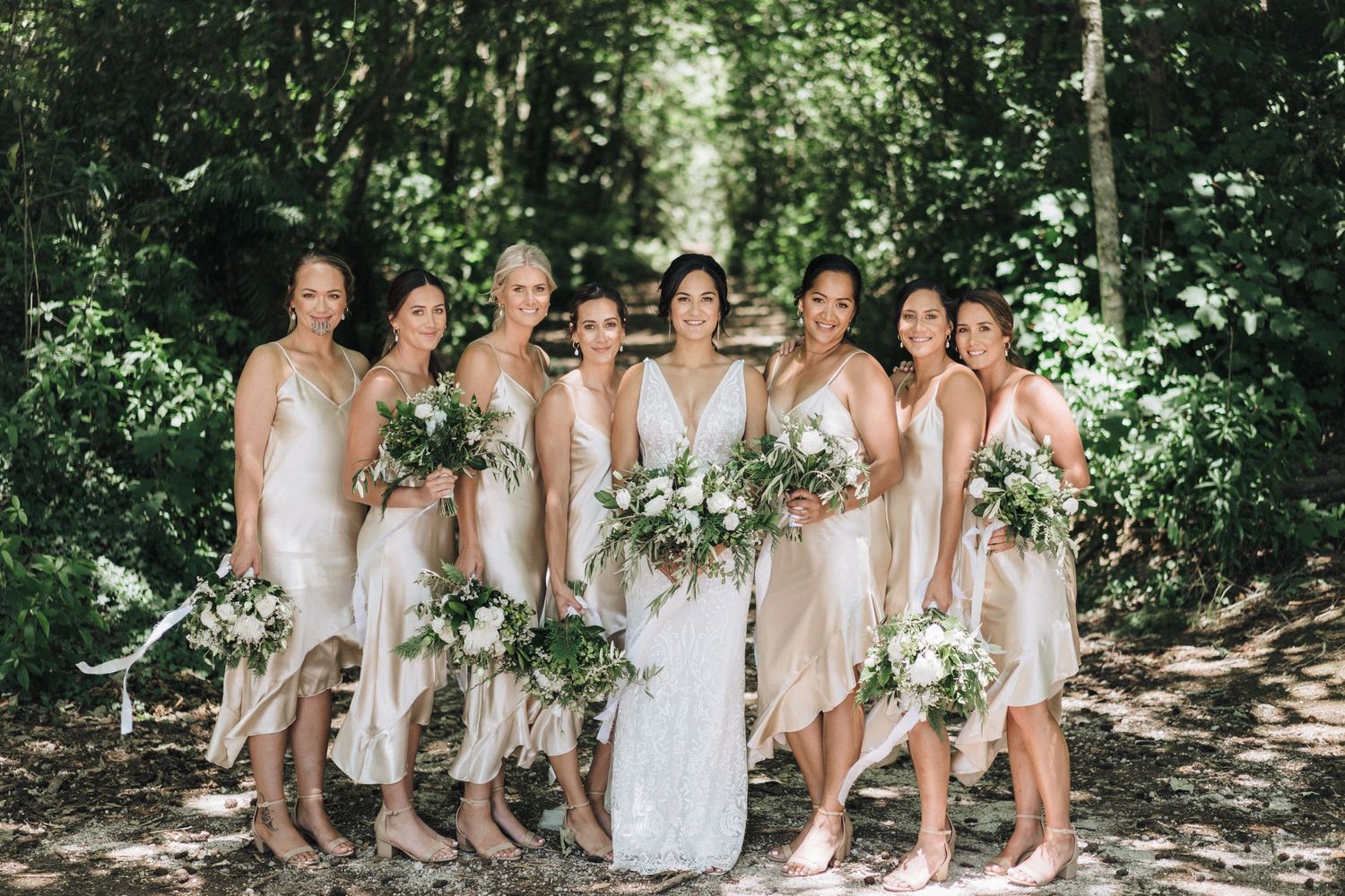 Elegant bridal party in light blush dresses poses together in sunlit wooded setting with white bouquets.