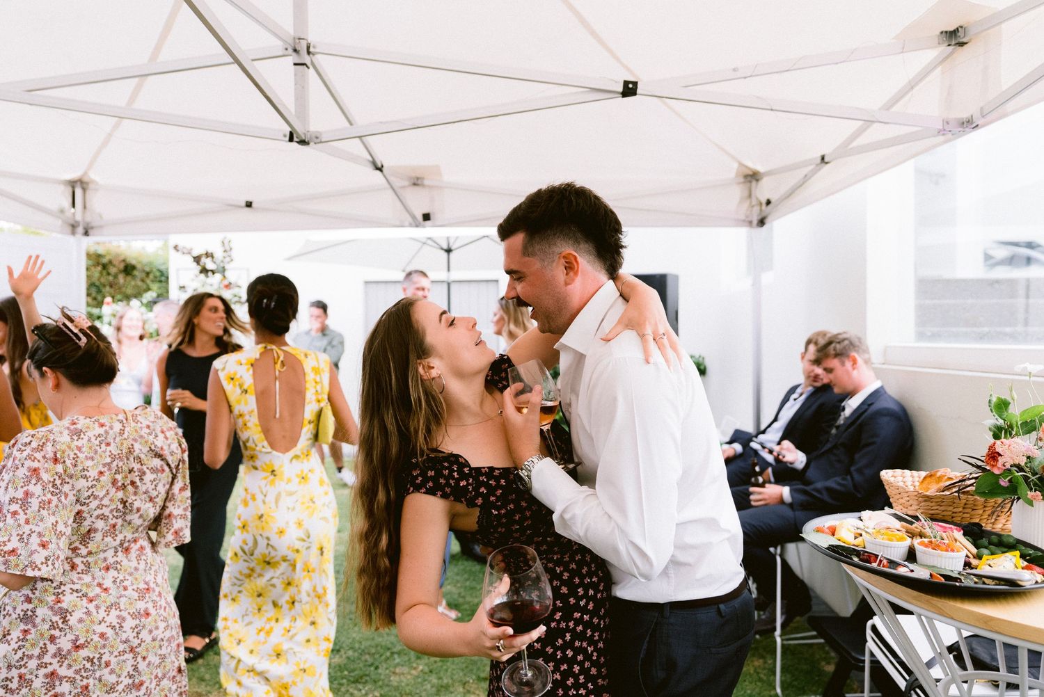 Wedding guests mingle and socialize under a white outdoor tent during a reception.