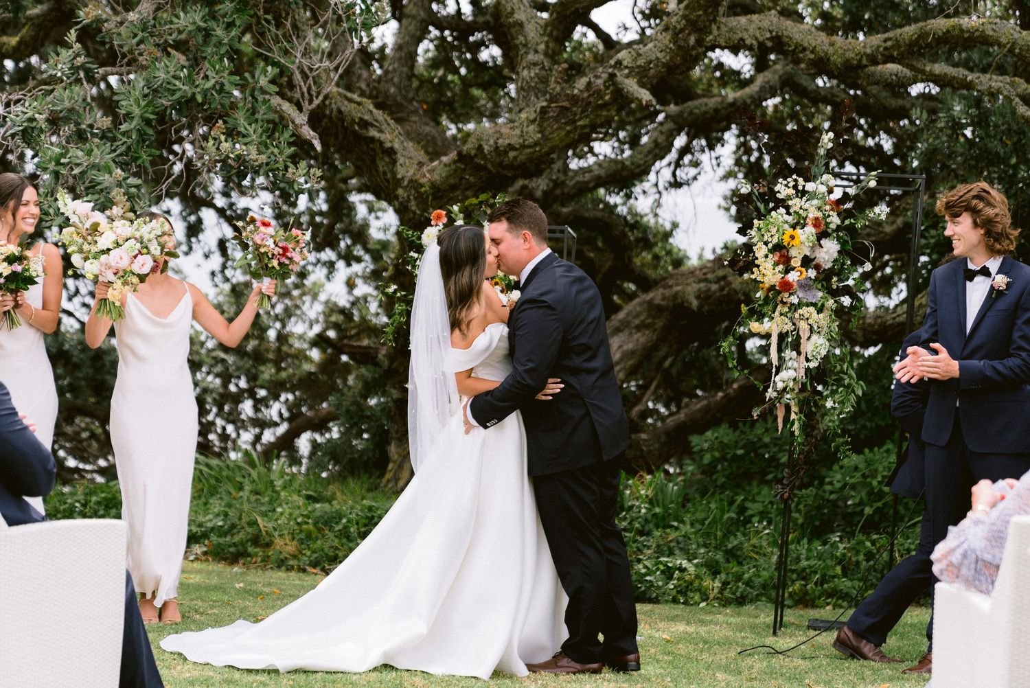 Wedding ceremony taking place under a large tree with floral decorations and gathered guests.