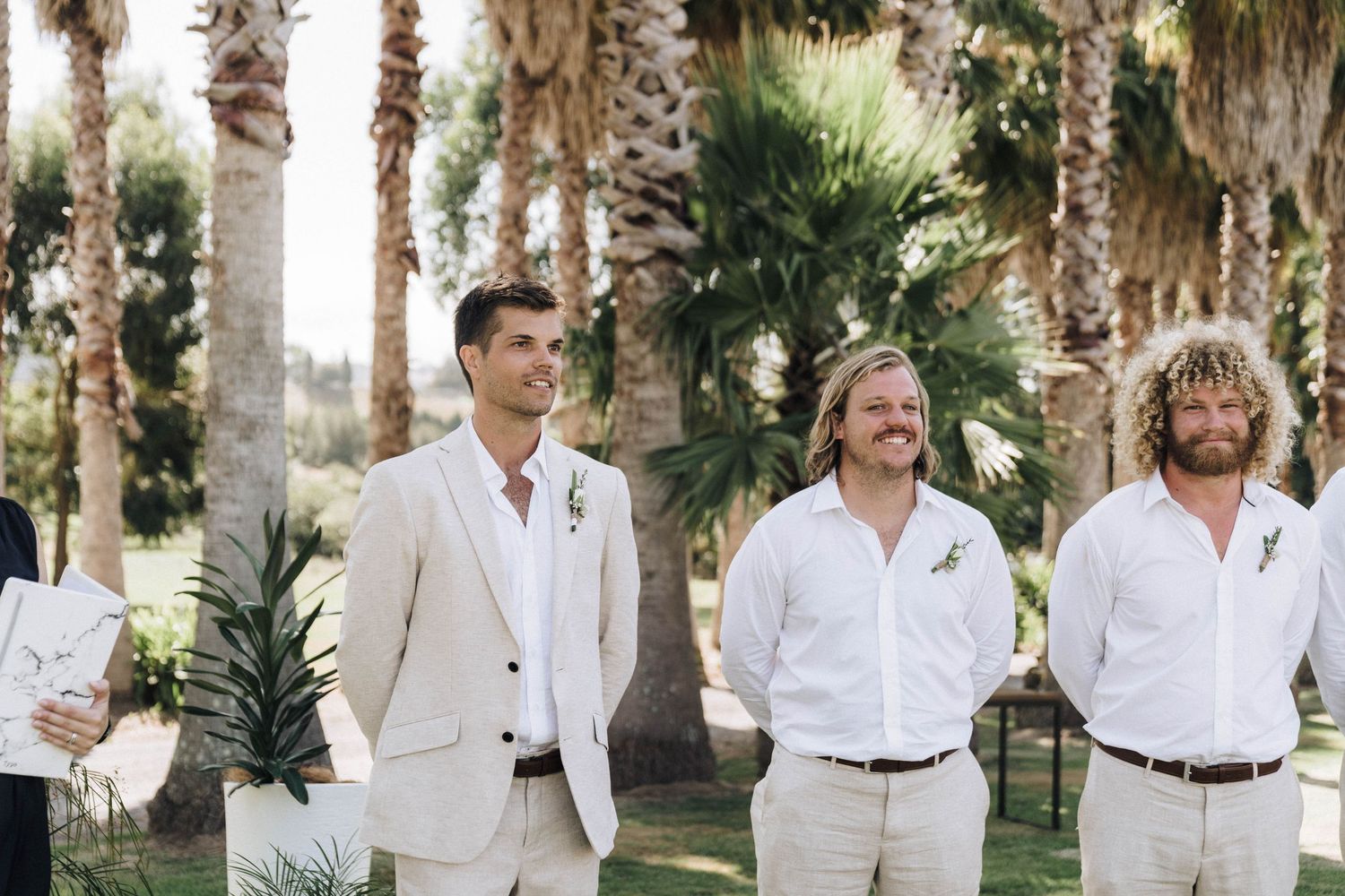 Wedding party members lined up outdoors among palm trees wearing light colored suits.