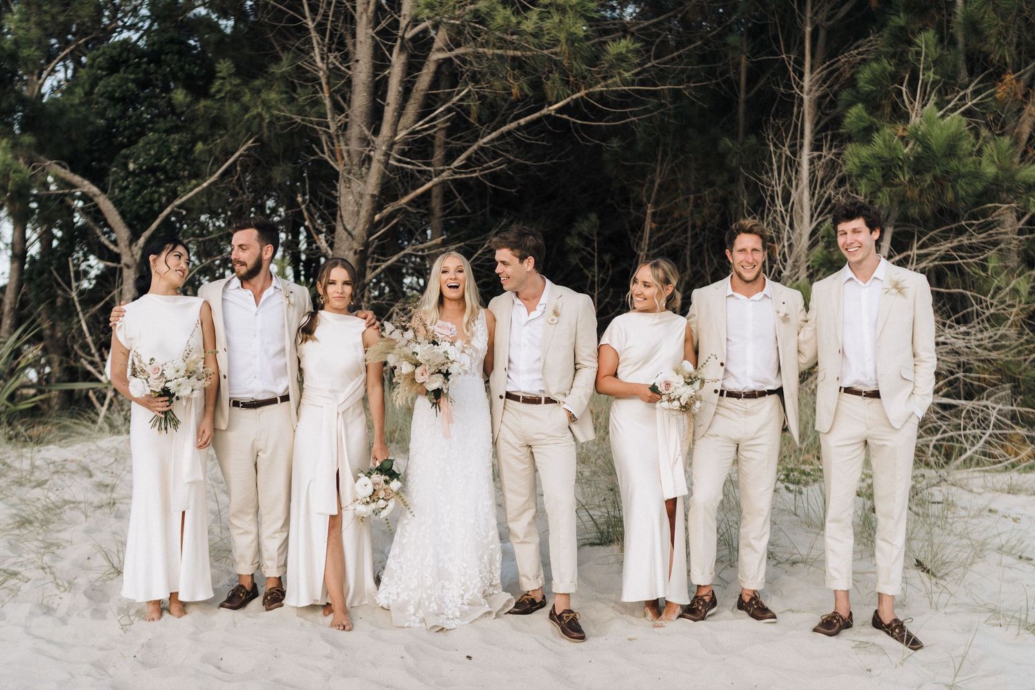 Wedding party in coordinated cream and beige outfits posing together on a beach.