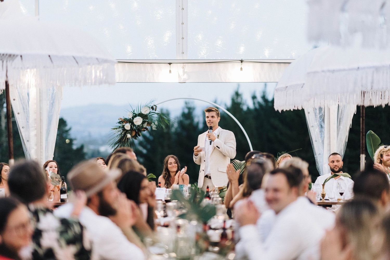 Guests seated at decorated outdoor wedding tables listen to toasts under white drapes and string lights.