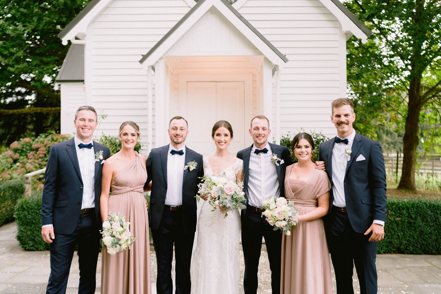 Wedding party in formal attire with black tuxedos and blush pink dresses poses together outside white chapel.