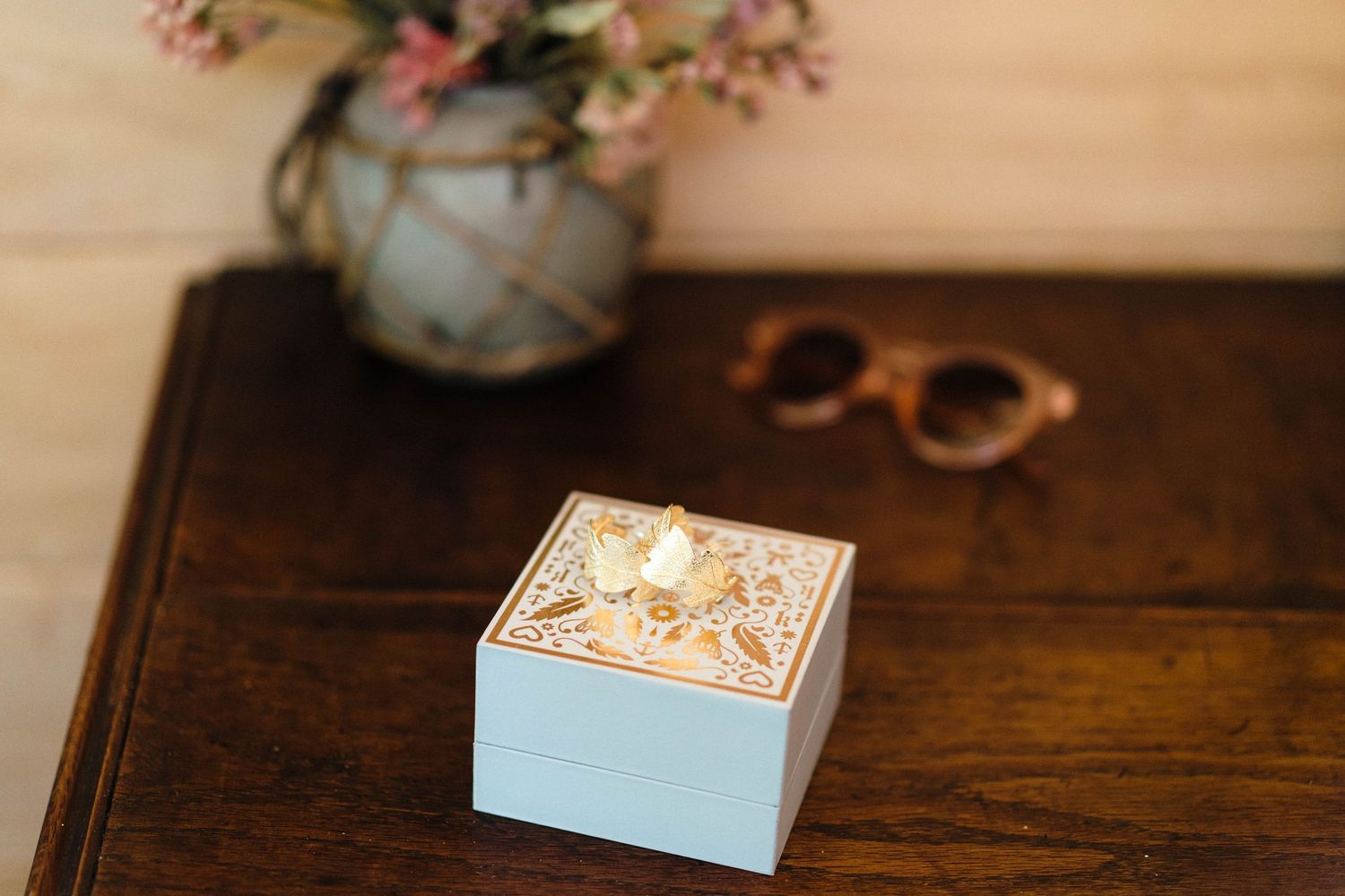 Decorative blue box and sunglasses on wooden table with floral arrangement in vase.