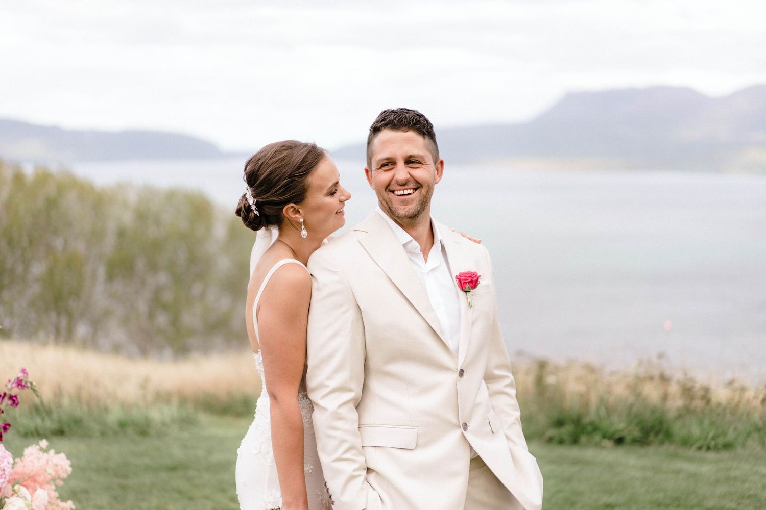 Couple in wedding attire pose on a hilltop with a scenic lake and mountains in the background.